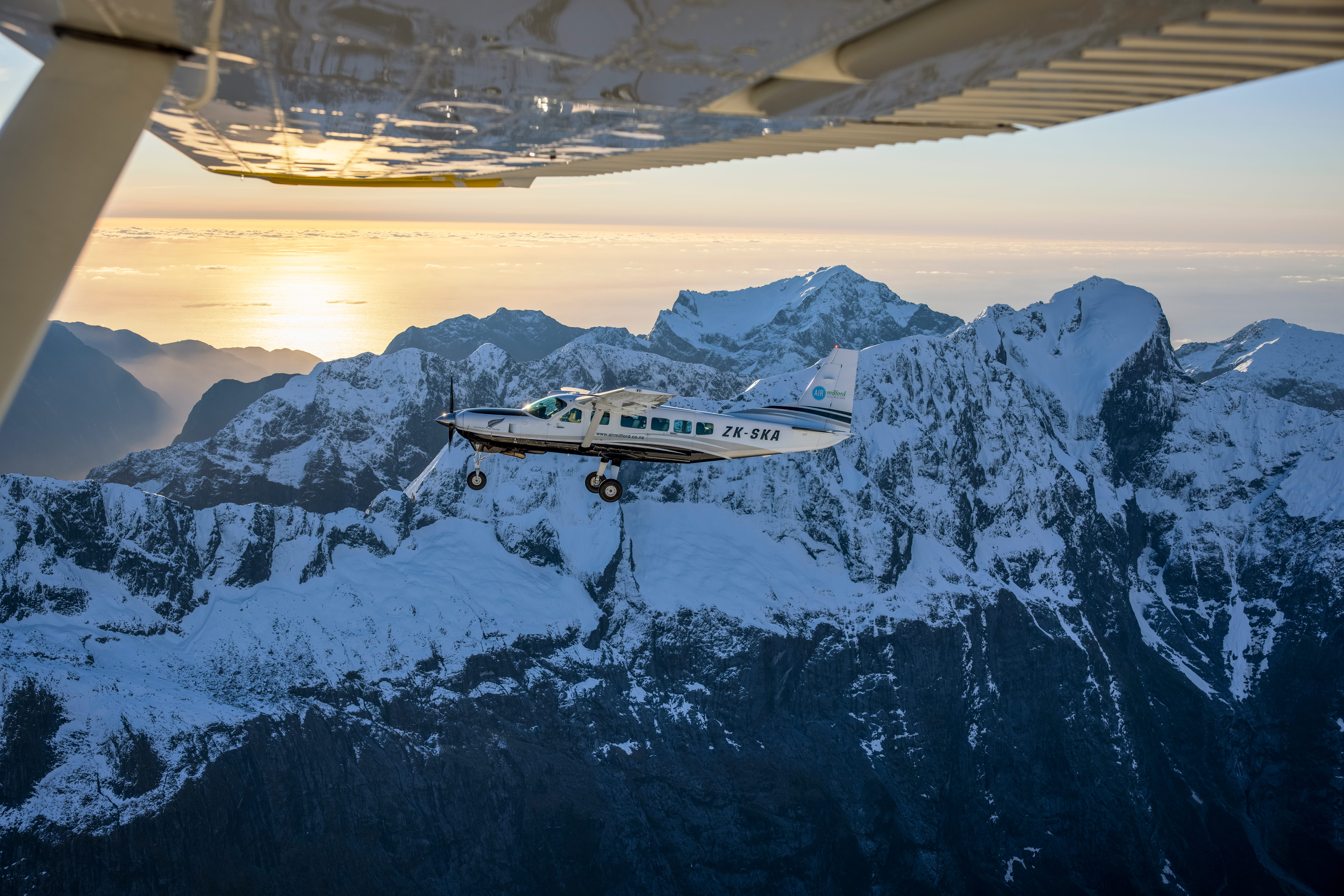A small scenic-flight aircraft flies over snow-covered mountain peaks at sunset.