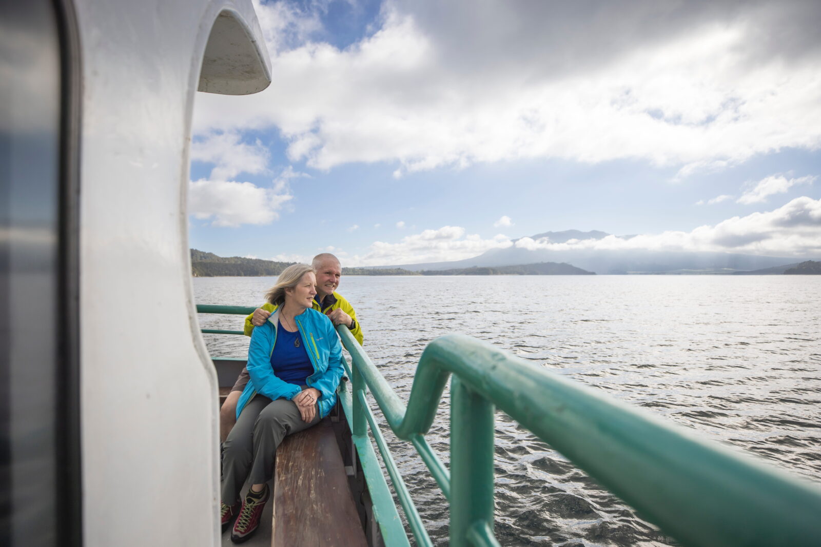 An older couple in rain jackets stand at a boat railing, looking out over sheltered, calm water with mountains in the distance.
