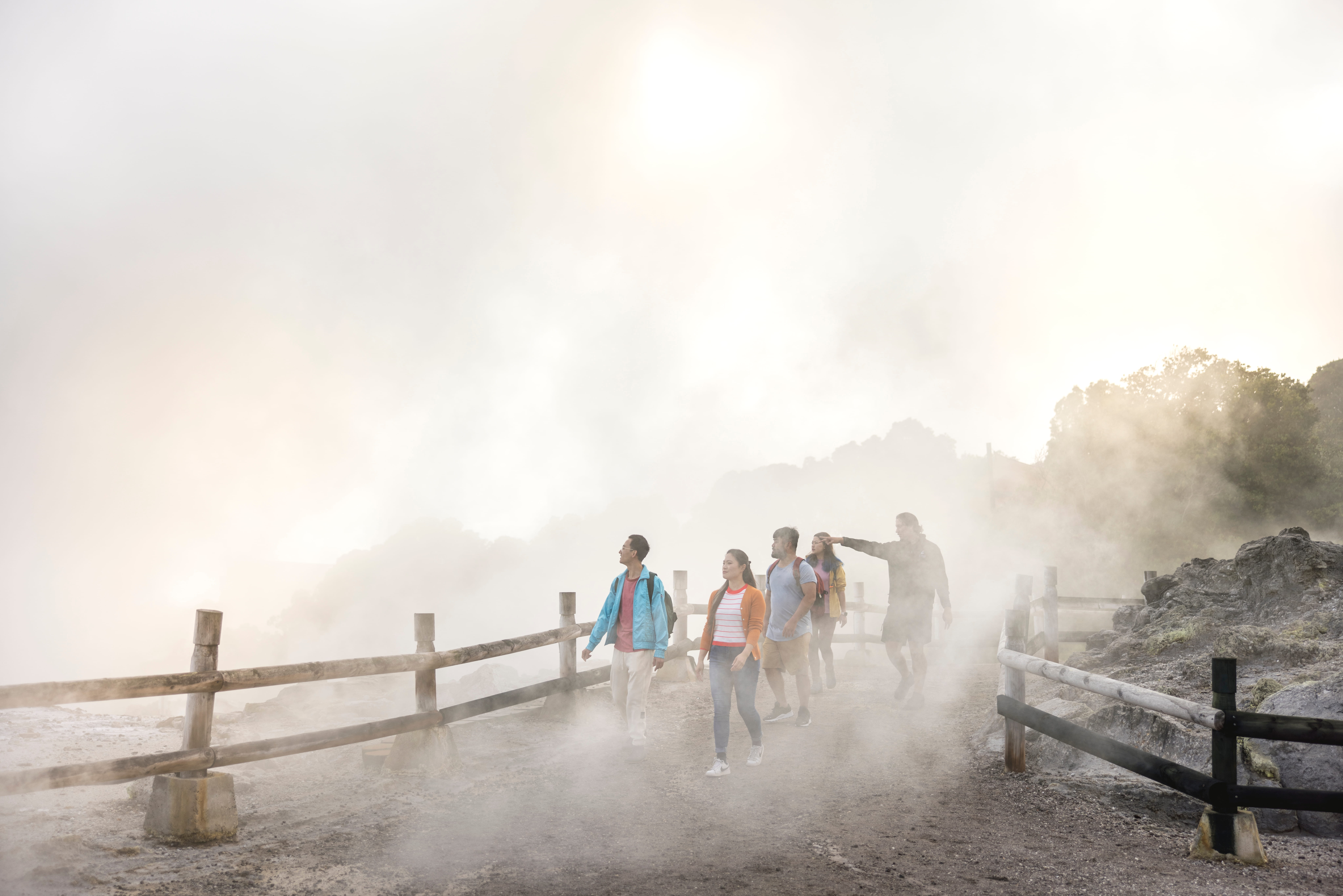 A small group of visitors walking along a wooden boardwalk through a steaming geothermal valley at sunrise, mist rising on either side.
