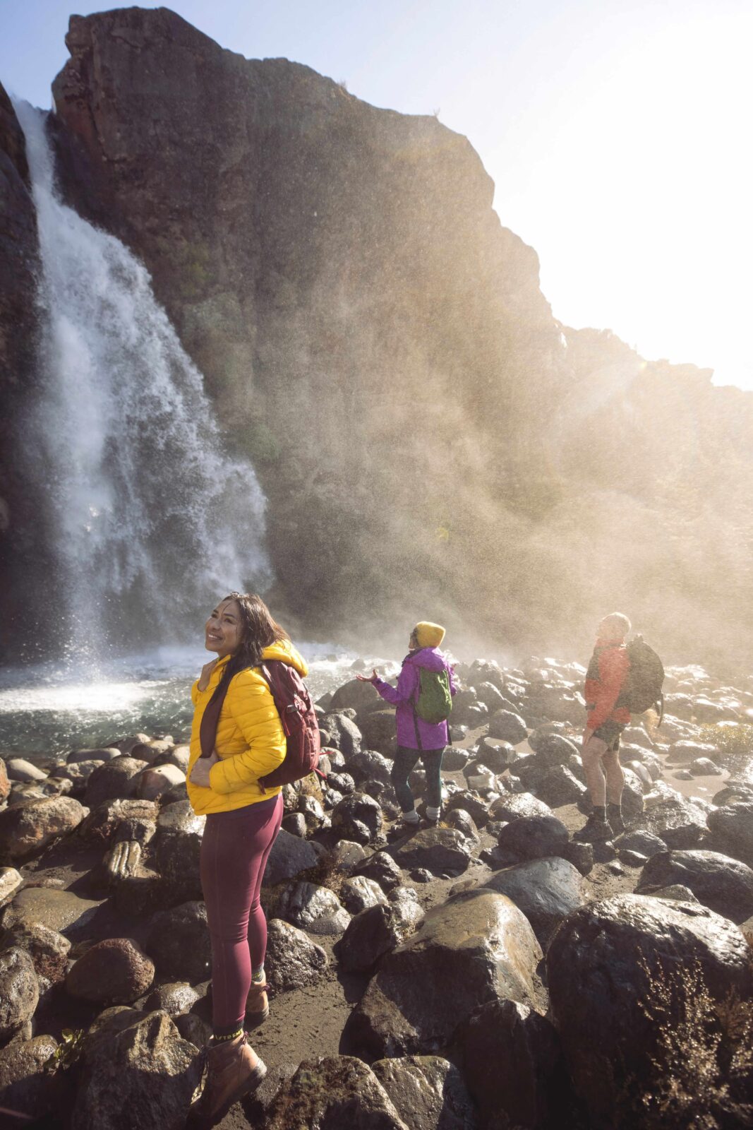 Three hikers in colorful jackets explore a boulder-strewn beach beneath a tall waterfall, backlit by hazy sunlight and mist.