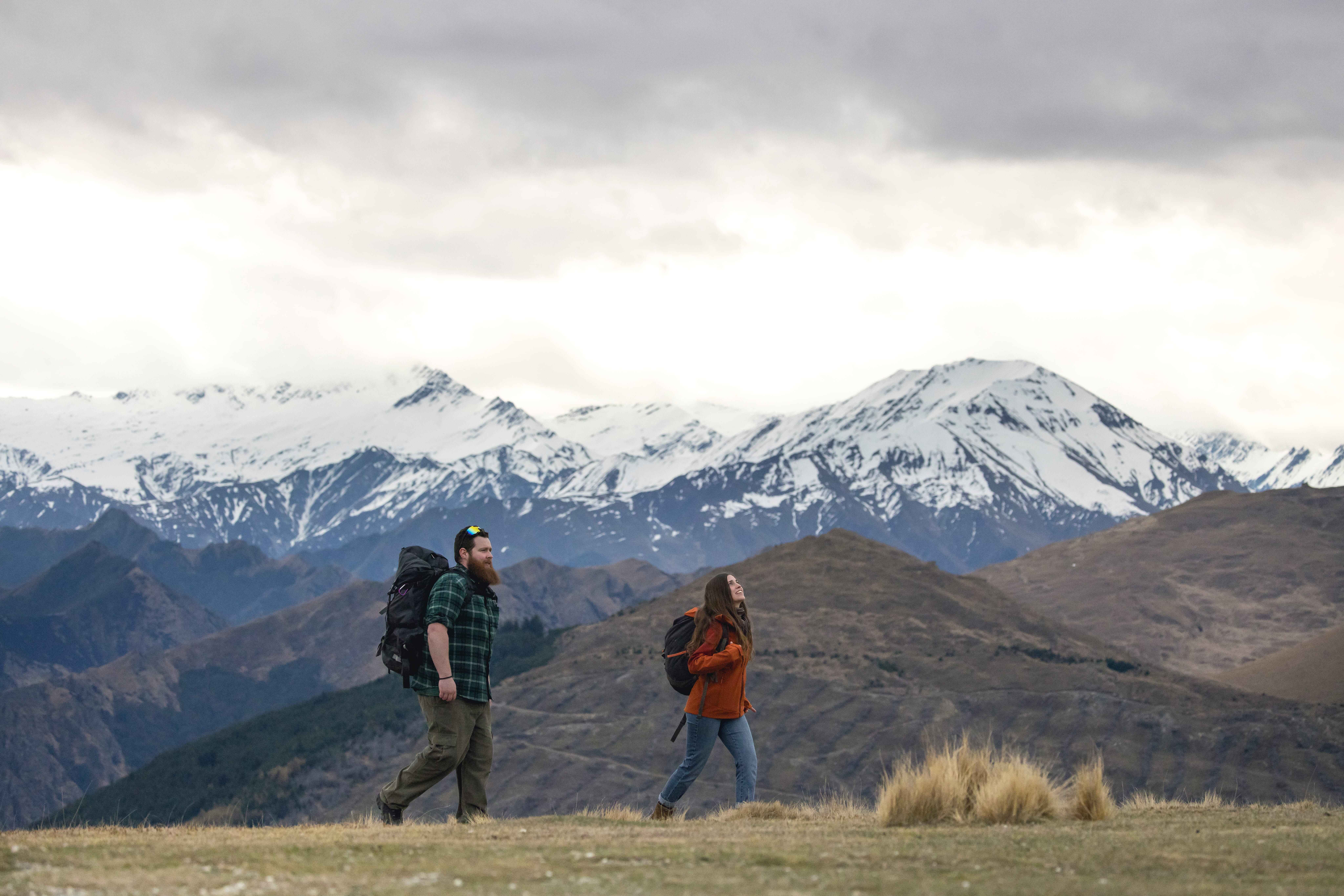  A couple hiking near Queenstown wearing backpacks. The Southern Alps tower in the background.