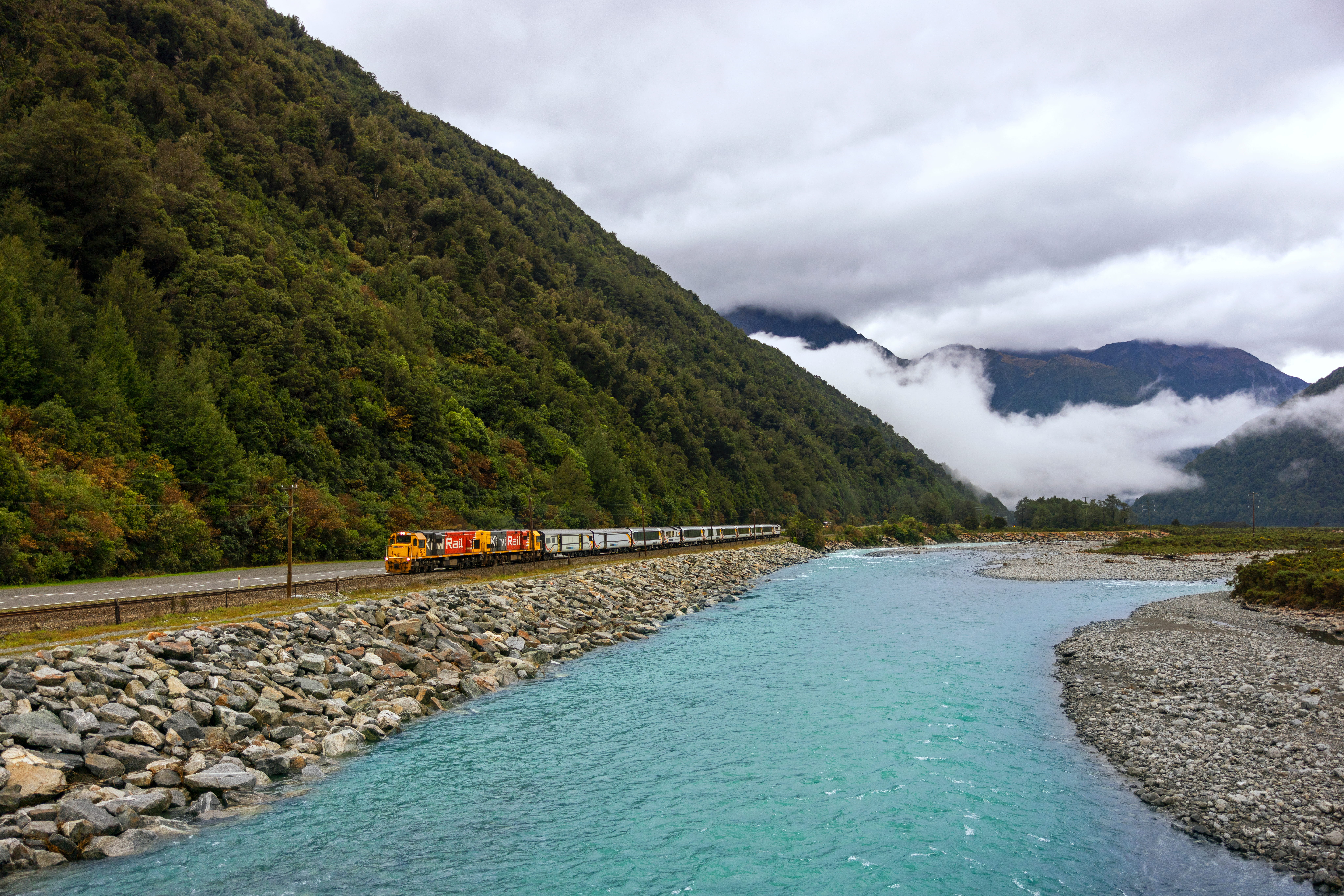 The TranzAlpine train travels along a rocky coastline beside turquoise water, with forested mountains rising behind.