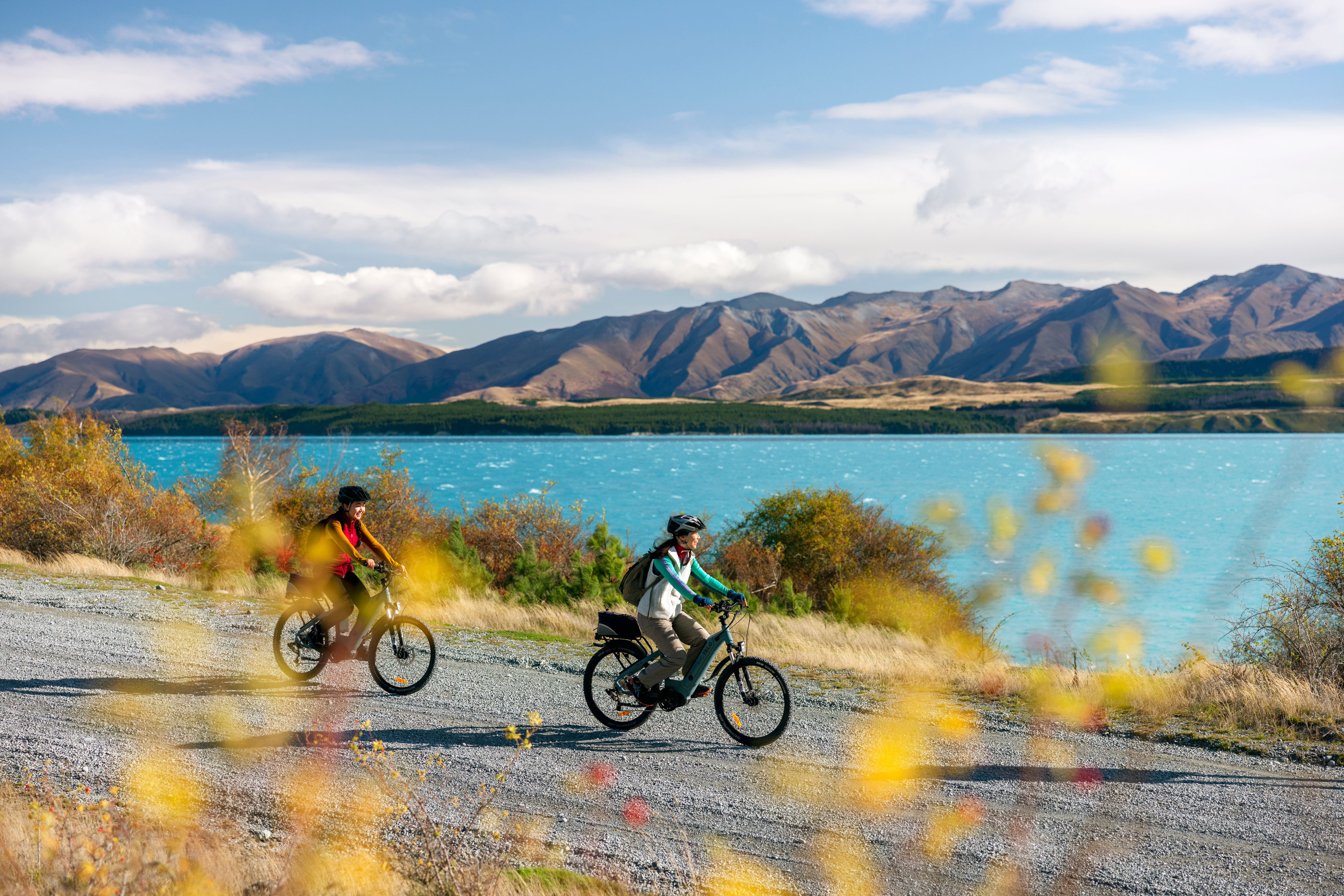 Two cyclists riding bikes along a gravel path beside a turquoise lake, with mountains in the background  