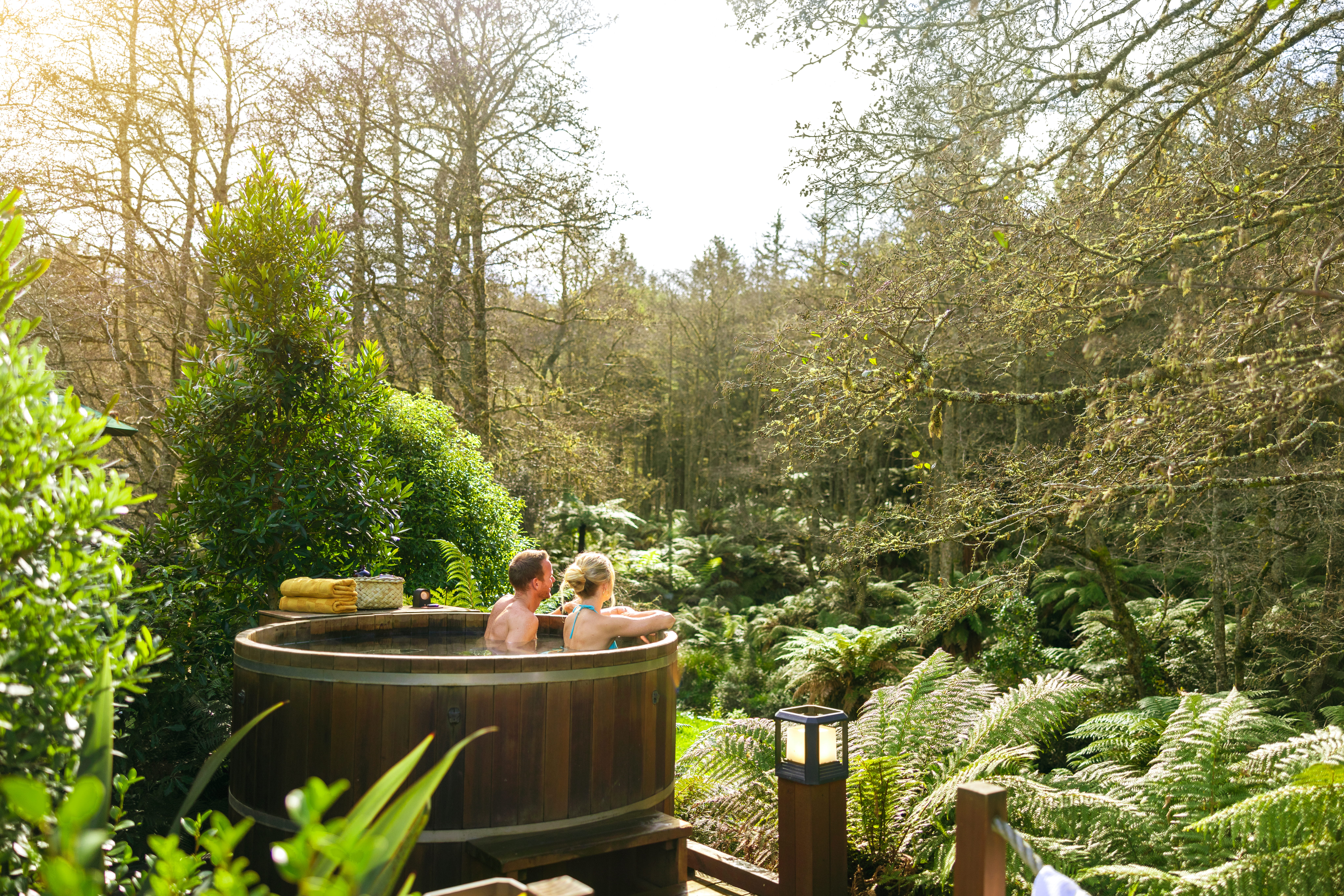 Two people relaxing in a wooden barrel hot tub on an outdoor deck, surrounded by lush garden greenery.