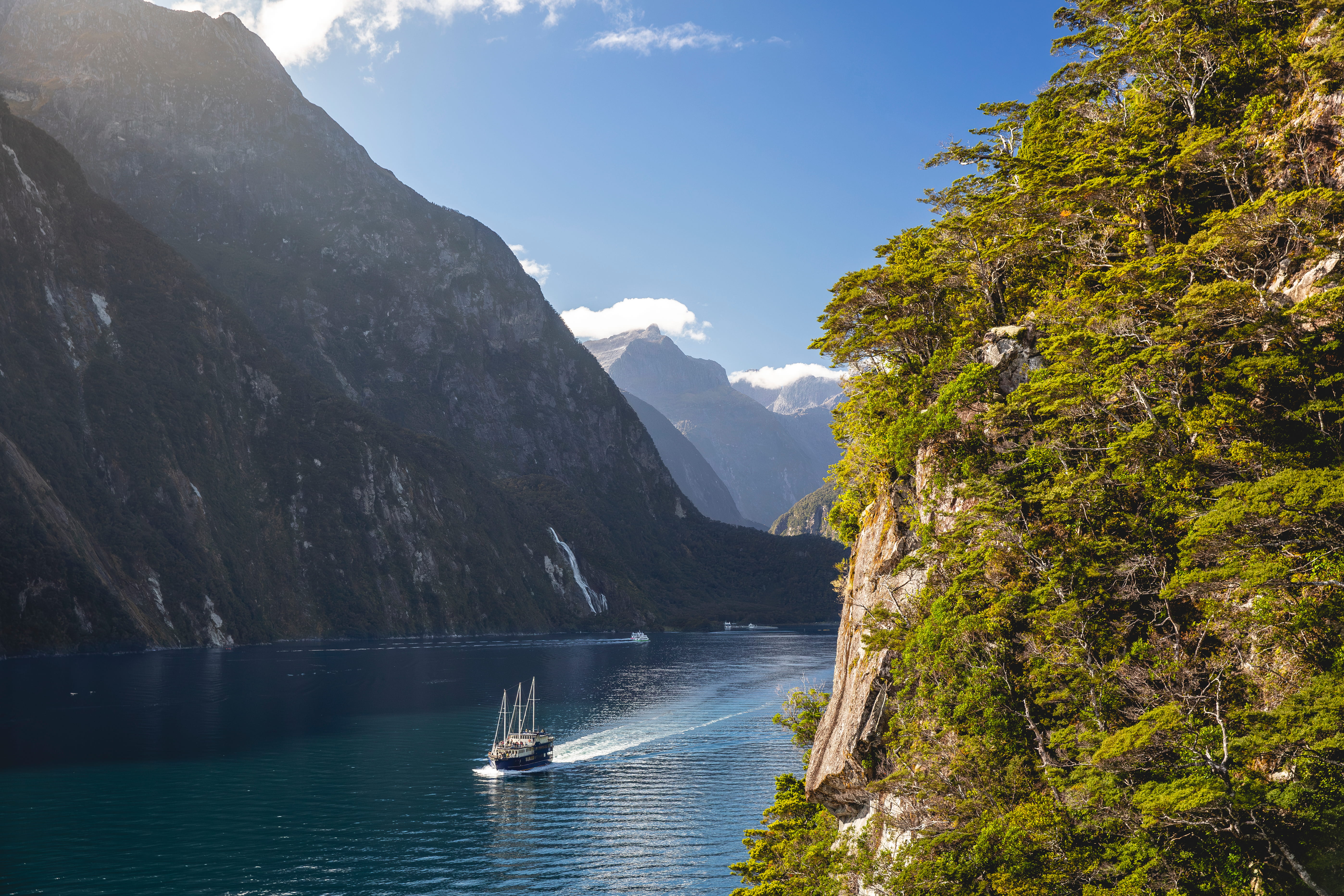 A tall-masted cruise boat travels through Milford Sound, a fjord with steep forested cliffs, waterfalls, and cloud-wrapped mountain peaks.