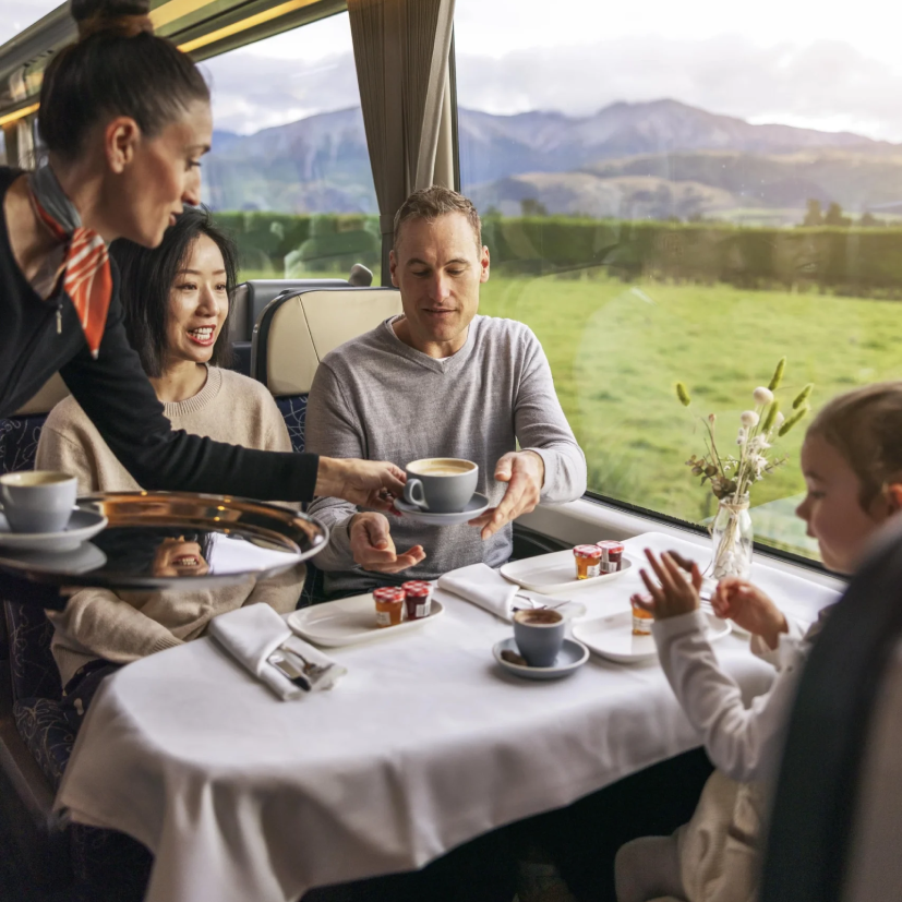 A family enjoys refreshments at a table while riding a scenic train through mountain landscapes.