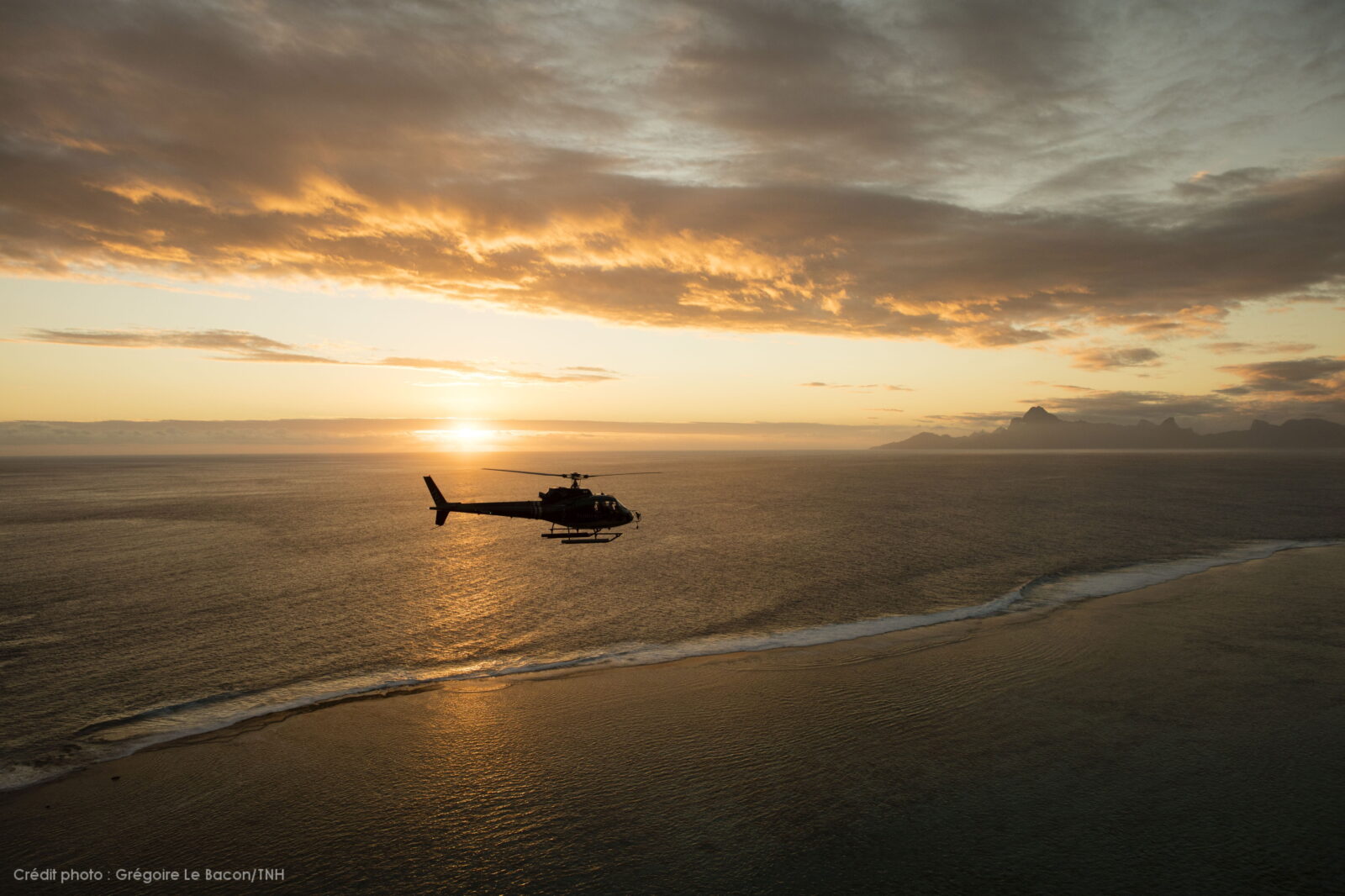 A helicopter flying along a coastline at sunset, silhouetted against the sky.
