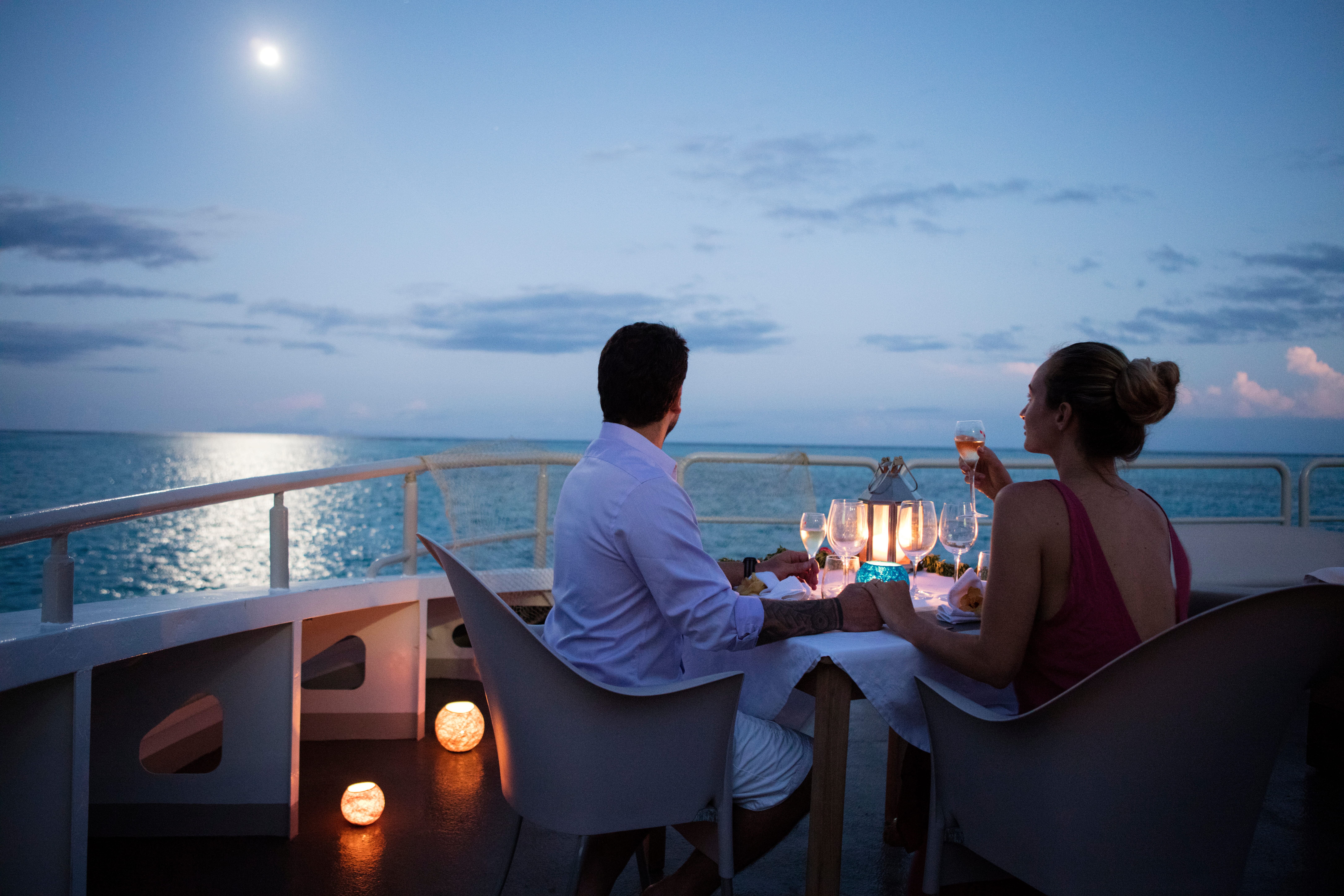 A couple having a candlelit dinner on a boat deck at dusk, overlooking the ocean.