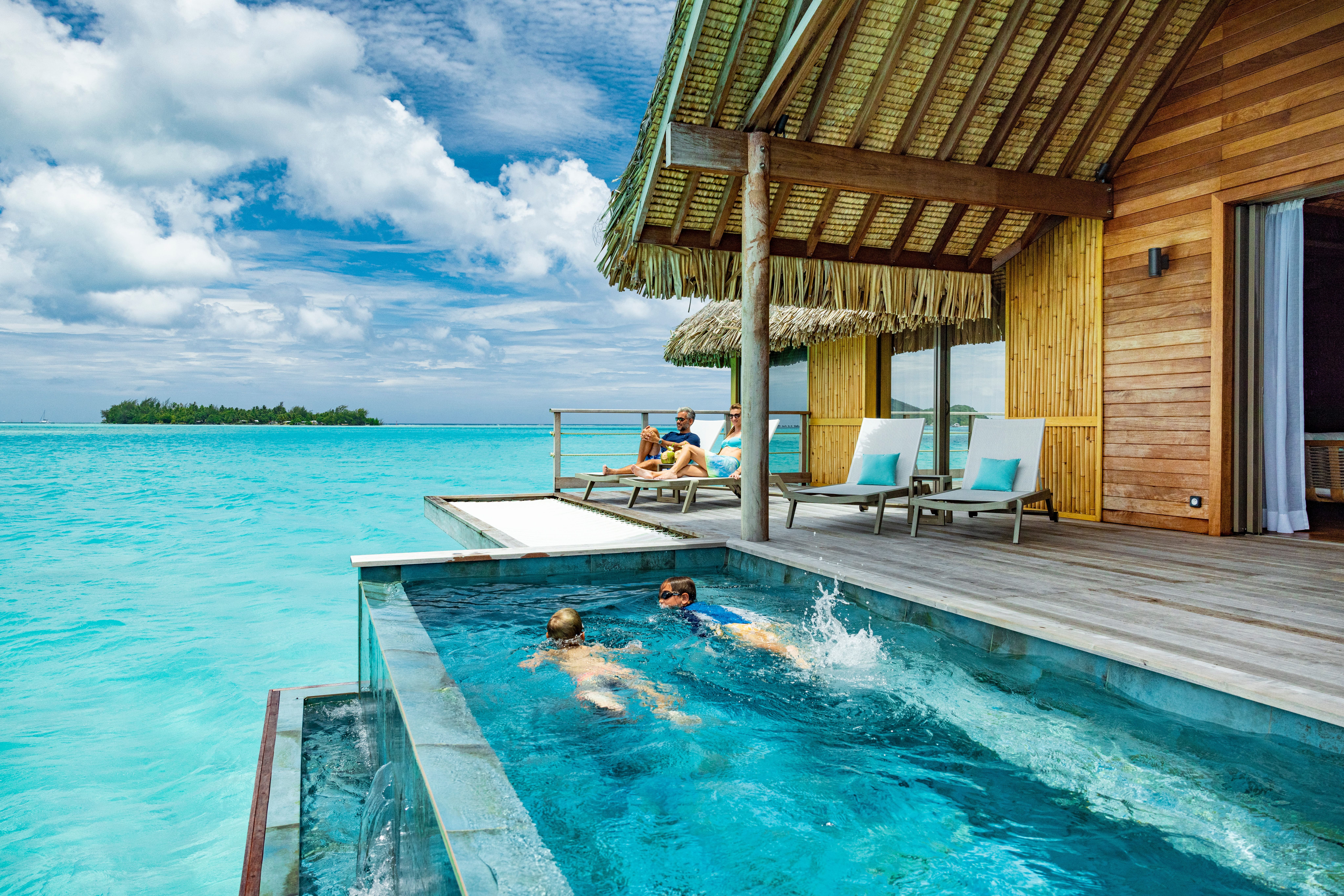 Two children swim in a private overwater plunge pool while adults relax on loungers under a thatched-roof deck, with a small island visible across the lagoon.
