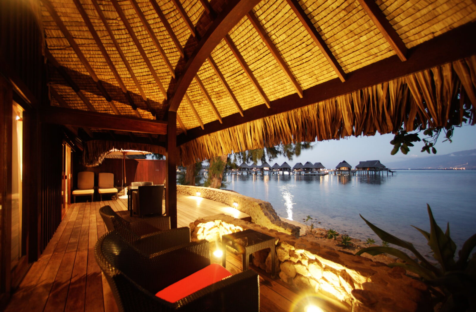 A thatched-roof villa lounge at dusk, overlooking overwater bungalows along the lagoon.