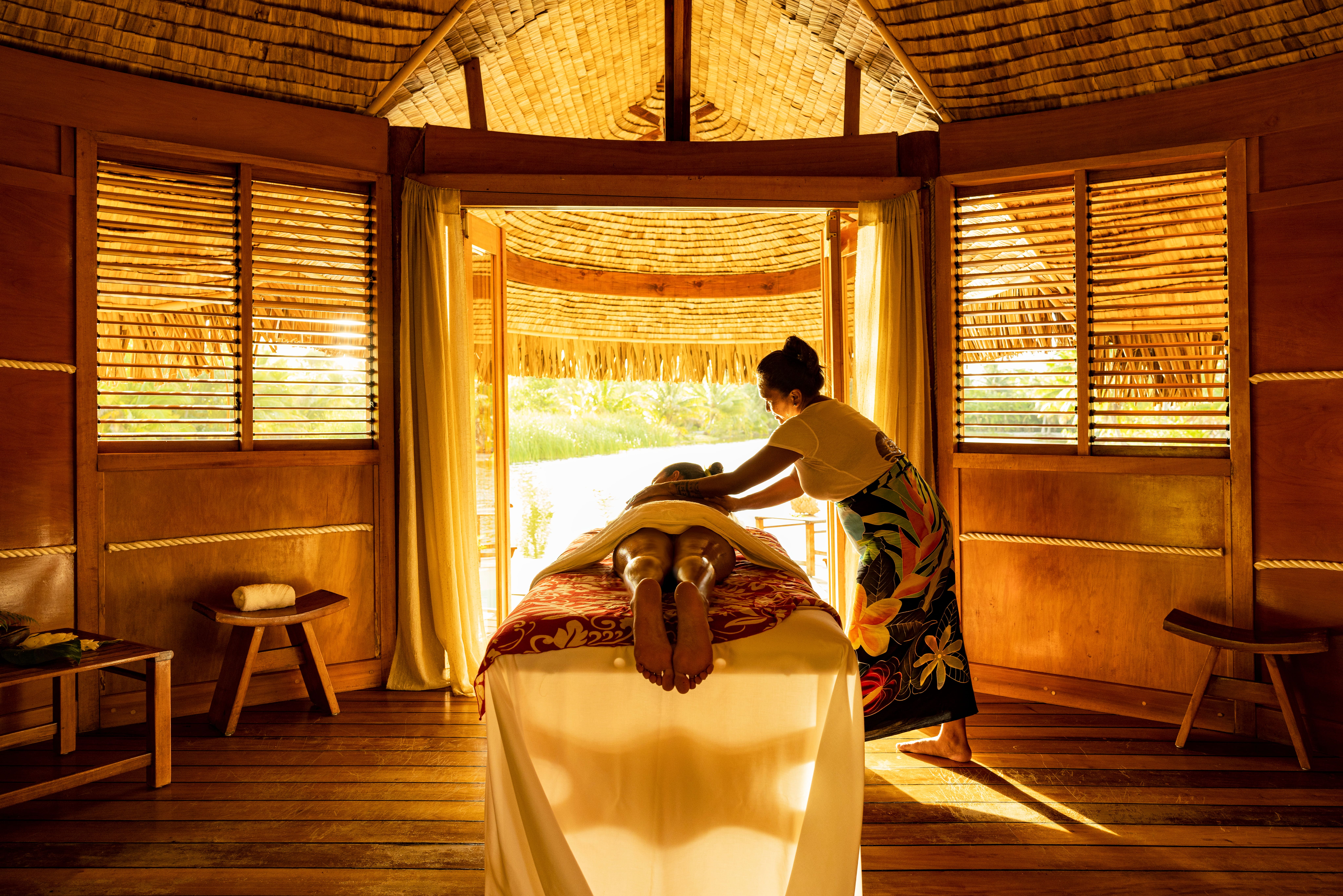 A guest receives a massage on a table inside an open-air overwater bure, with the ocean visible through the doorway.