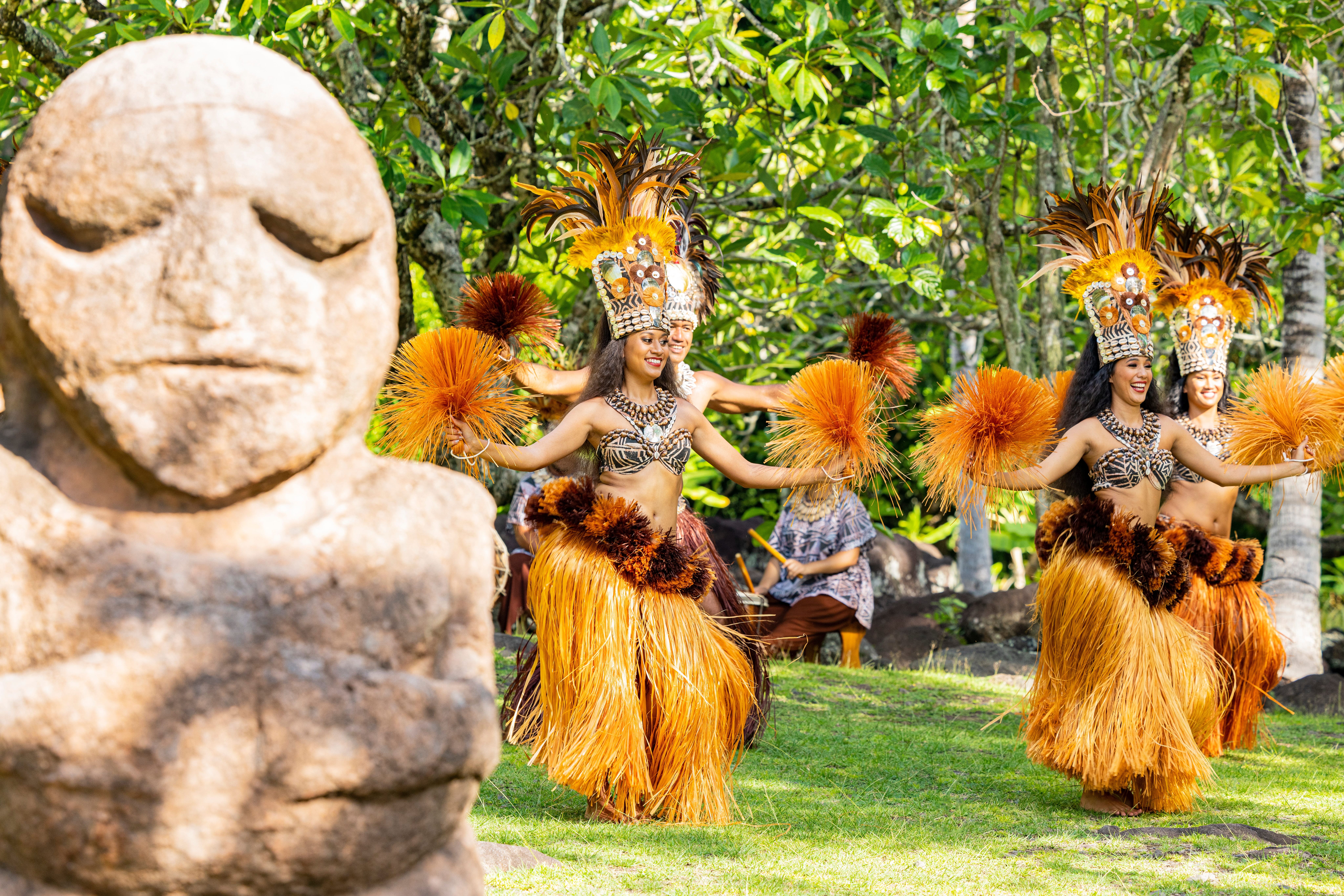 A carved stone tiki statue stands in the foreground while Polynesian dancers in feathered headdresses and grass skirts perform behind it.