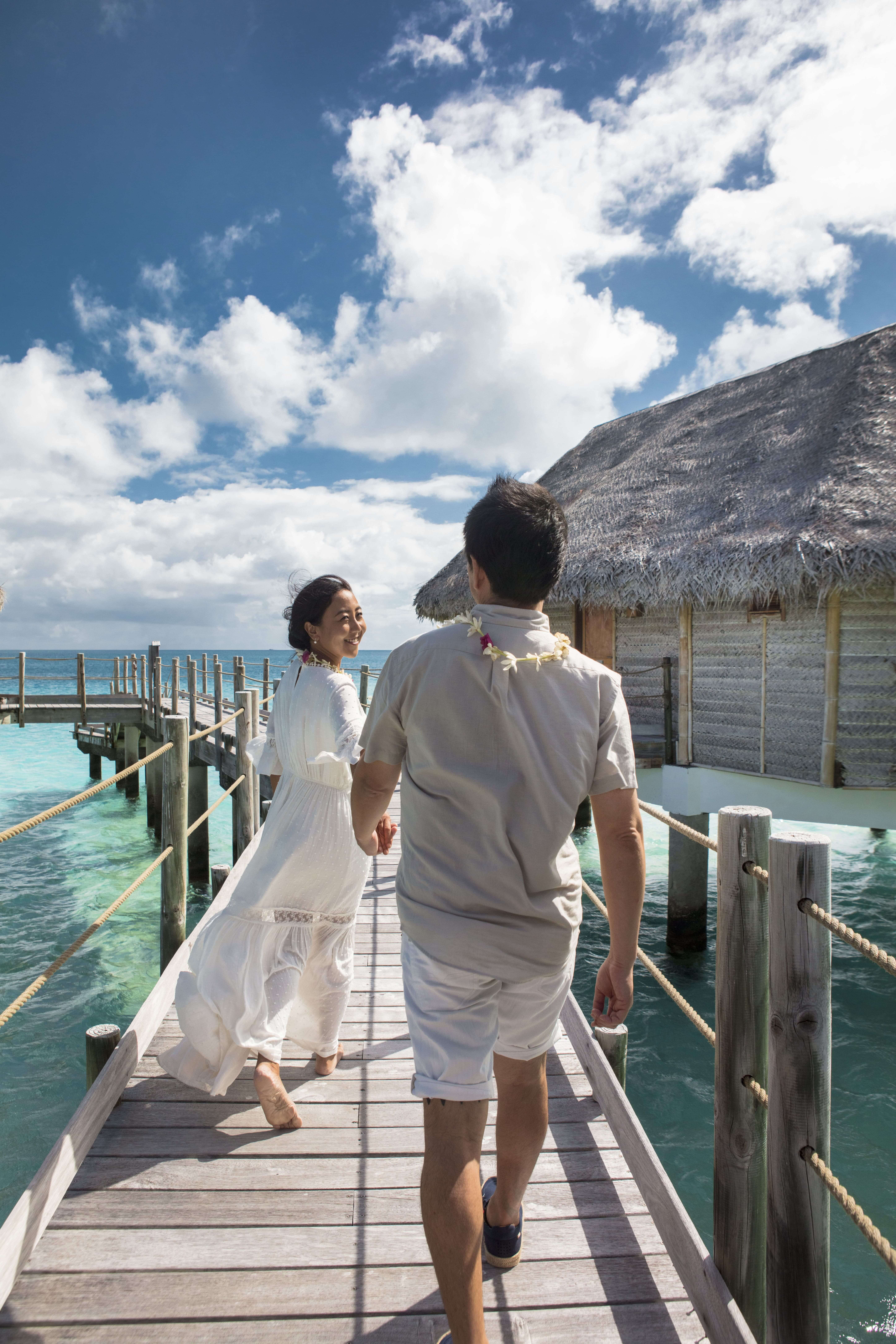 A couple in white walk hand in hand along a wooden overwater walkway toward a thatched-roof bungalow.