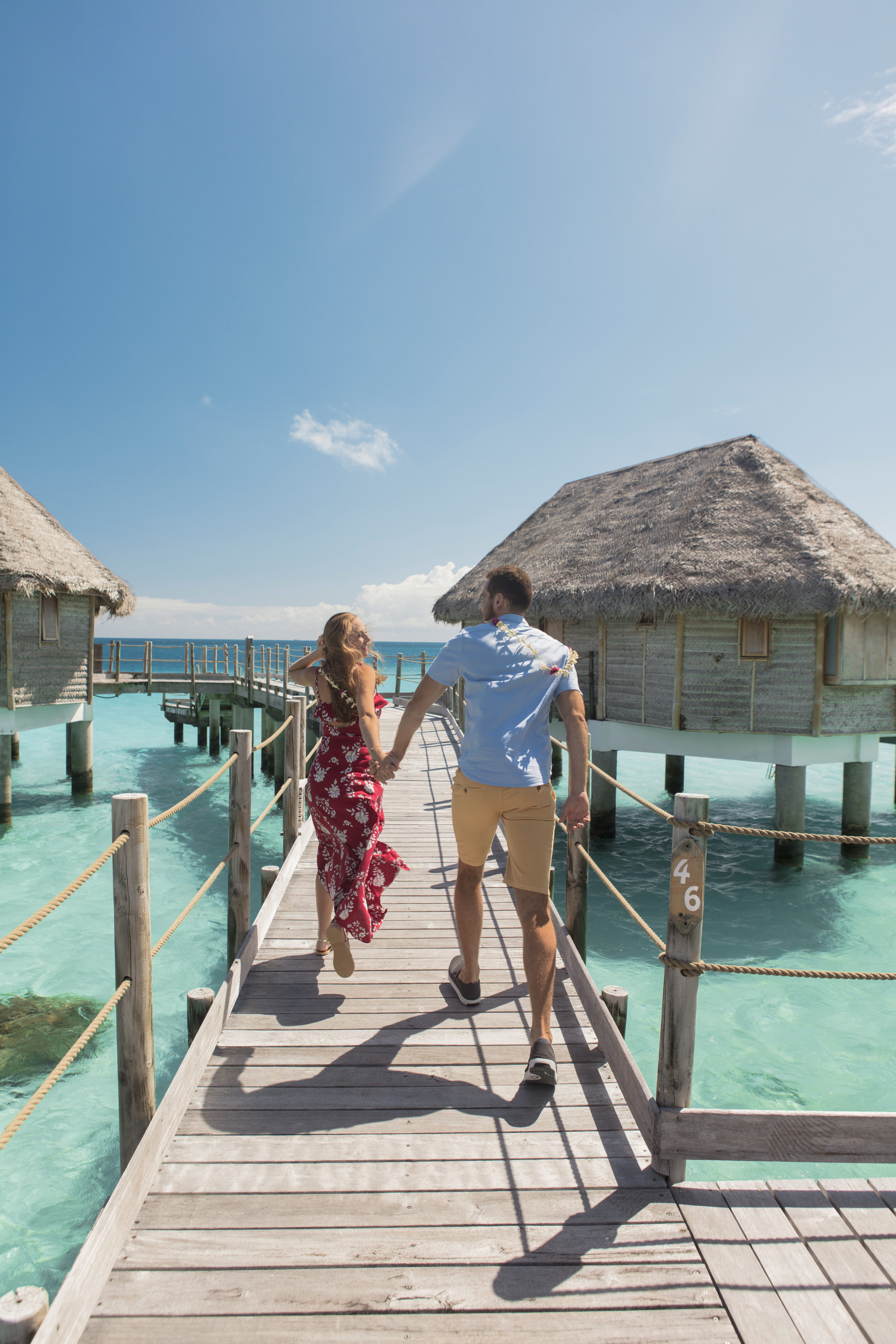 Happy couple walks hand in hand down a boardwalk, headed towards an overwater bungalow.