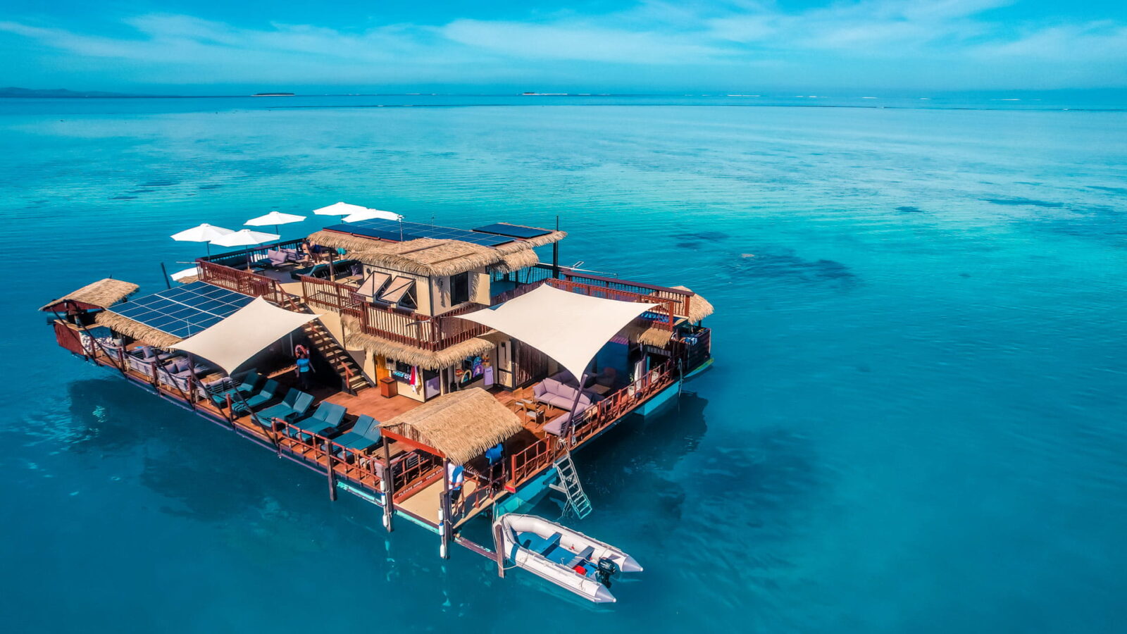 An aerial view of a floating overwater day club with thatched roofs and lounge decks, anchored in a turquoise lagoon with a speedboat moored alongside.