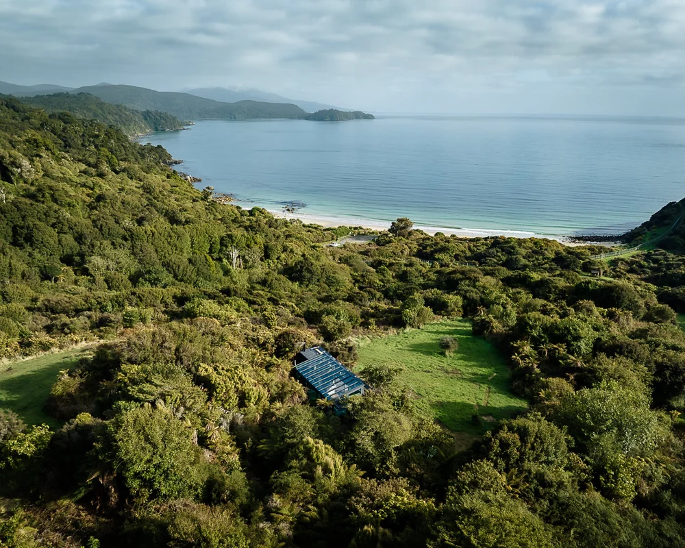 Aerial view of a solitary lodge with a dark roof tucked in a clearing amid dense native bush, overlooking the coastline in the distance.