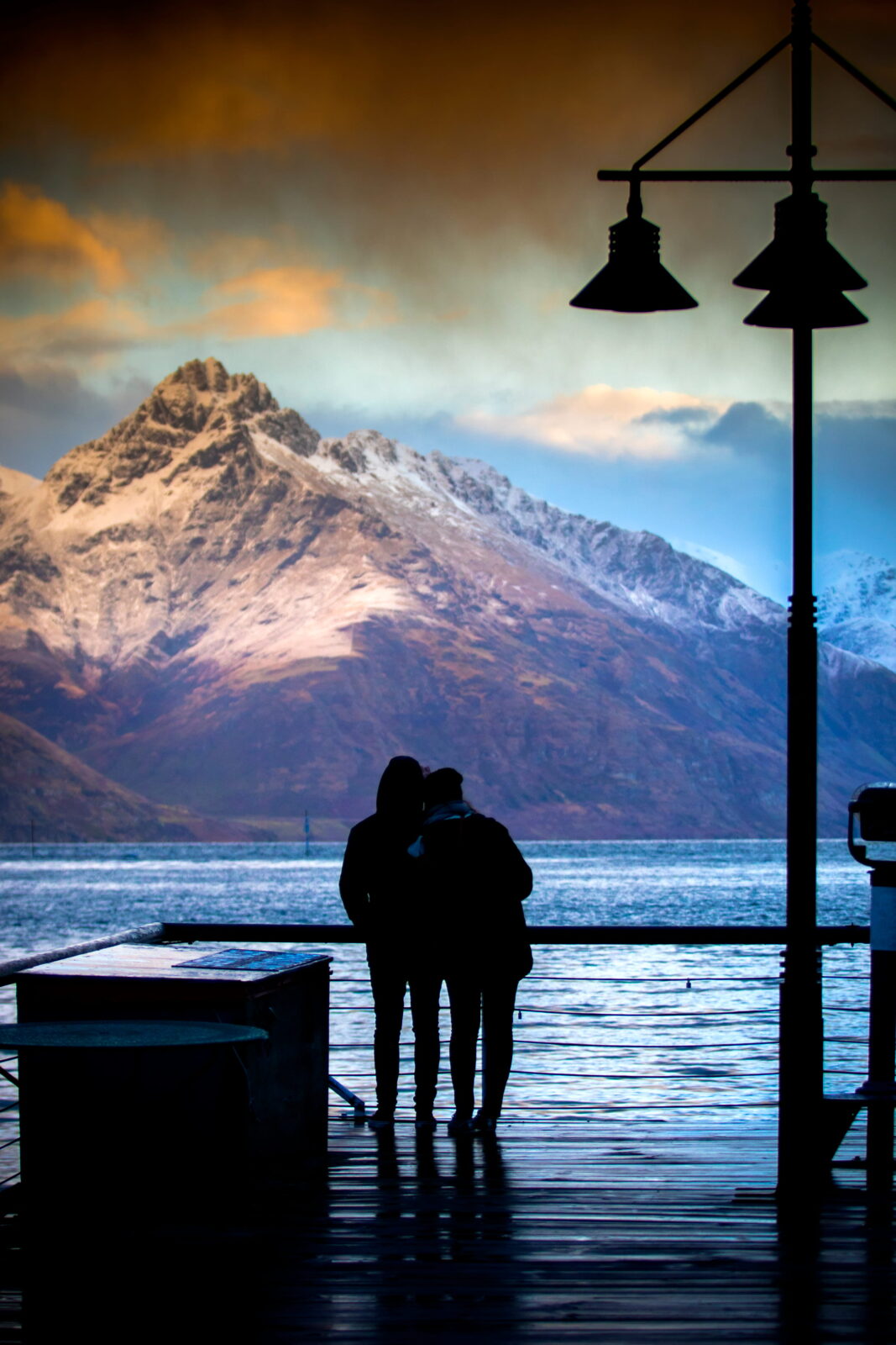 Silhouette of couples standing on Queenstown harbour pier against beautiful mountain scene.