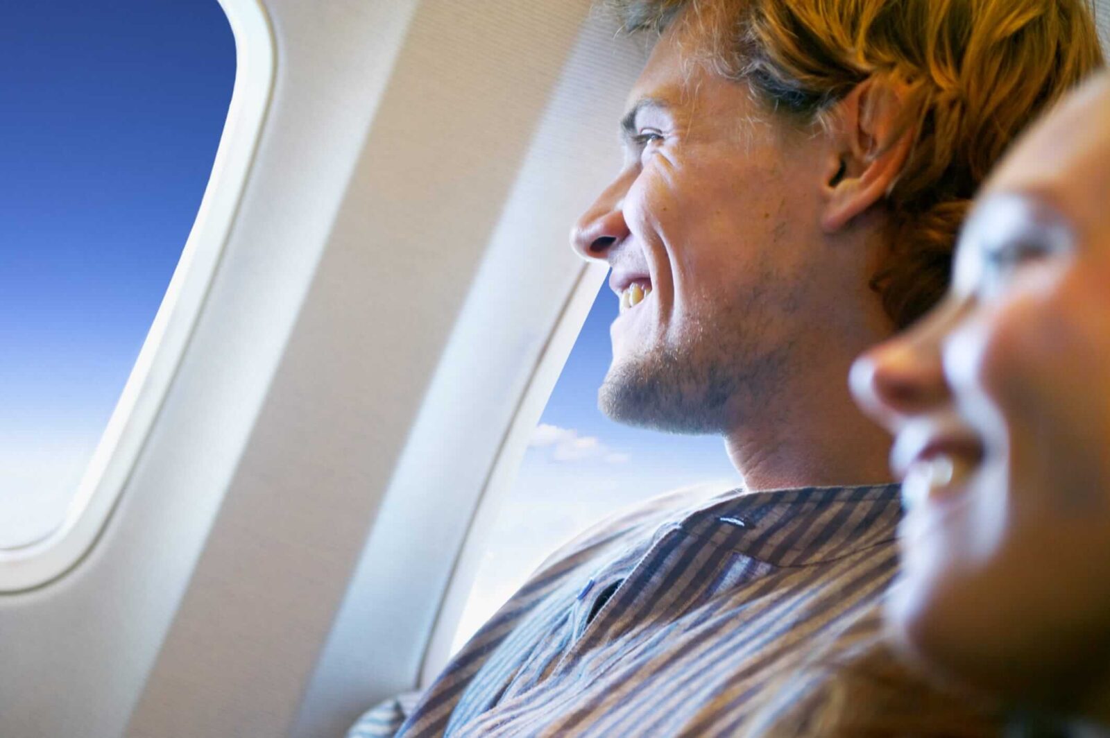 A passenger smiling while looking out an airplane window during a flight, with another passenger seated beside them.