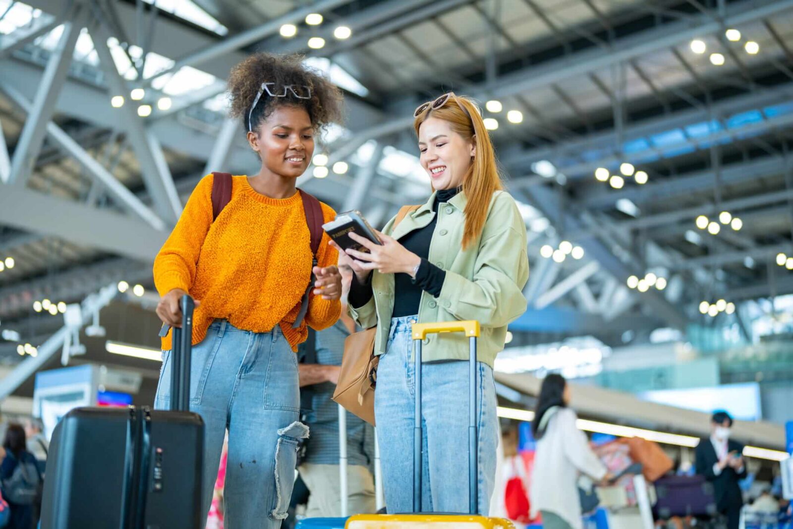 Two women travelers looking at a phone together in an airport terminal, one holding onto a suitcase