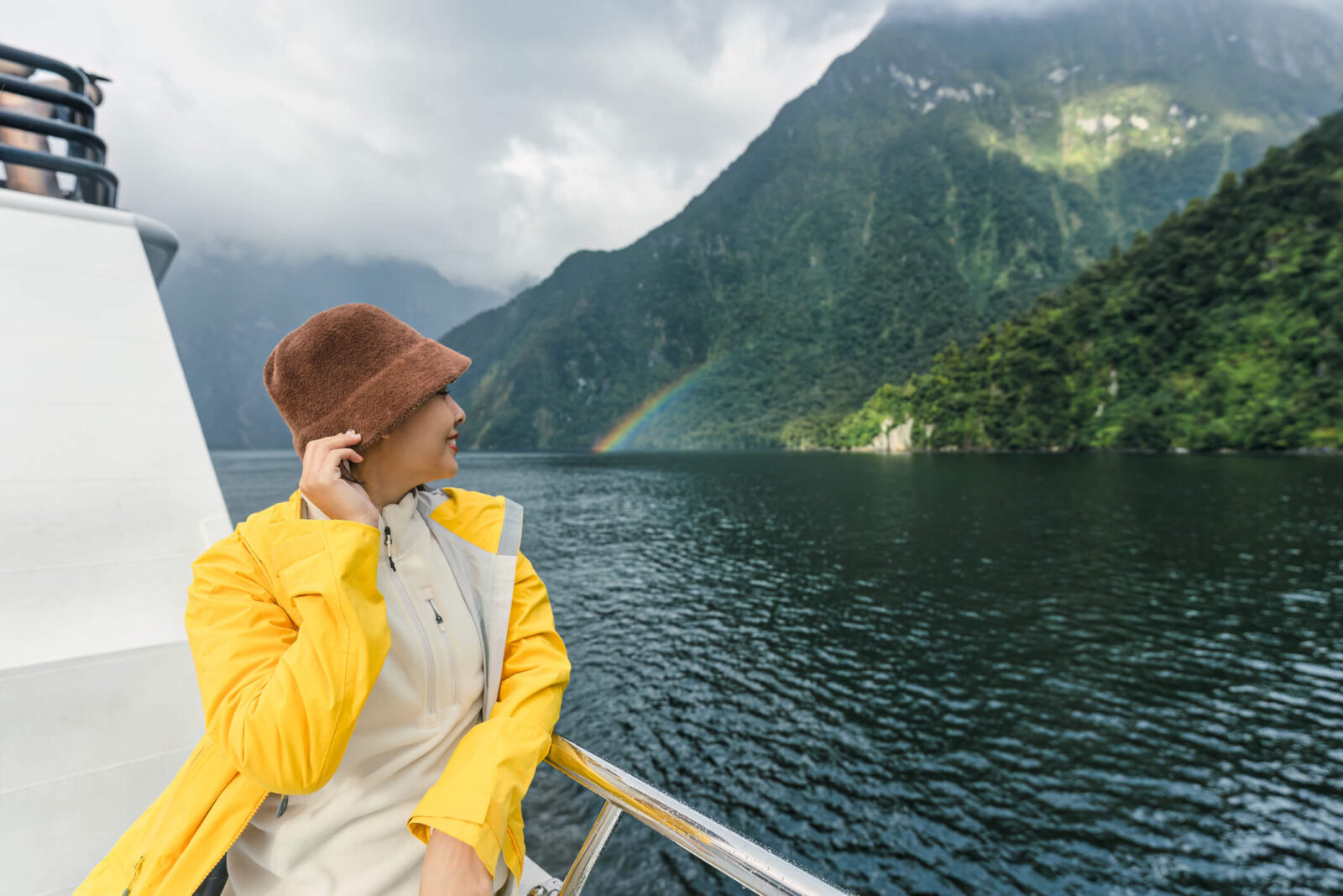 Asian woman wearing yellow jacket enjoying with mountain and rainbow on a ferry crusing in fjord on the lake at Milford Sound, New Zealand