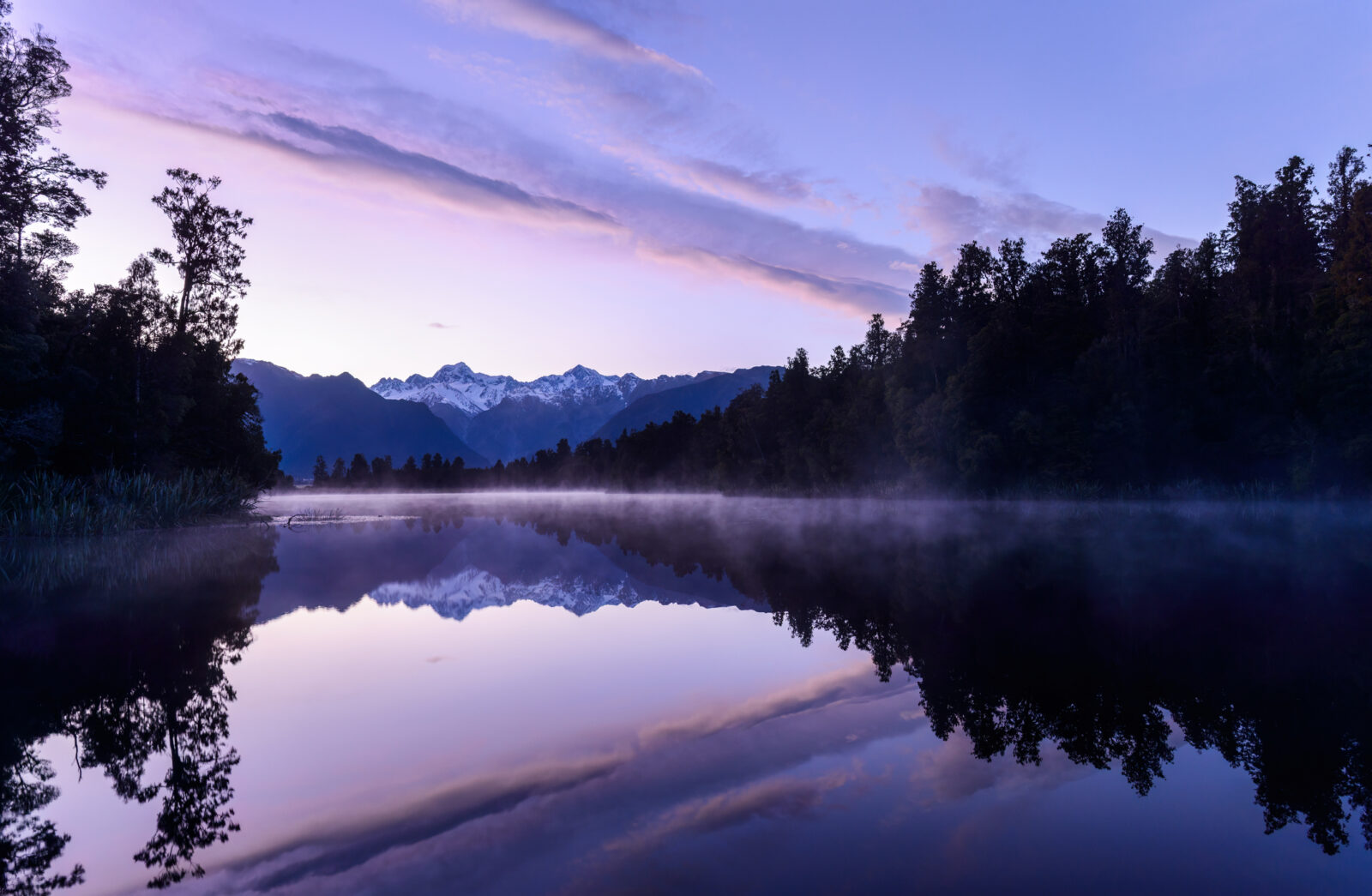 Dusk descends upon the glorious mirror lake that is Lake Matheson on New Zealand's South Island. In the distance we see the snow capped peaks of Mt Tasman, Mt Cook and the Southern Alps, all bathed in a magnificent Alpenglow, the last light of day.
