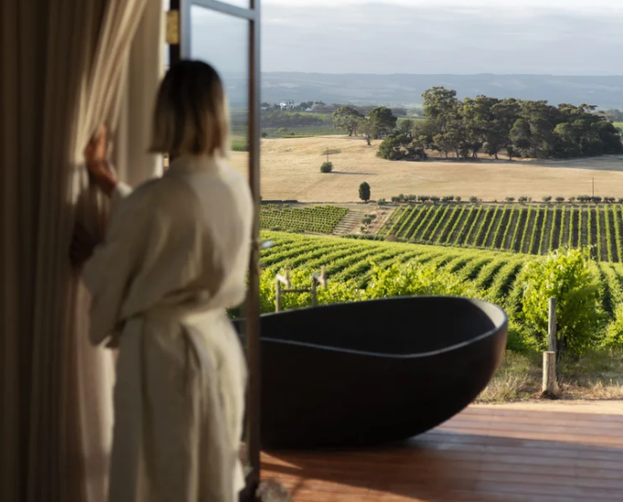 A person in a robe looks out over vineyard views beside a freestanding black bathtub on a private deck.