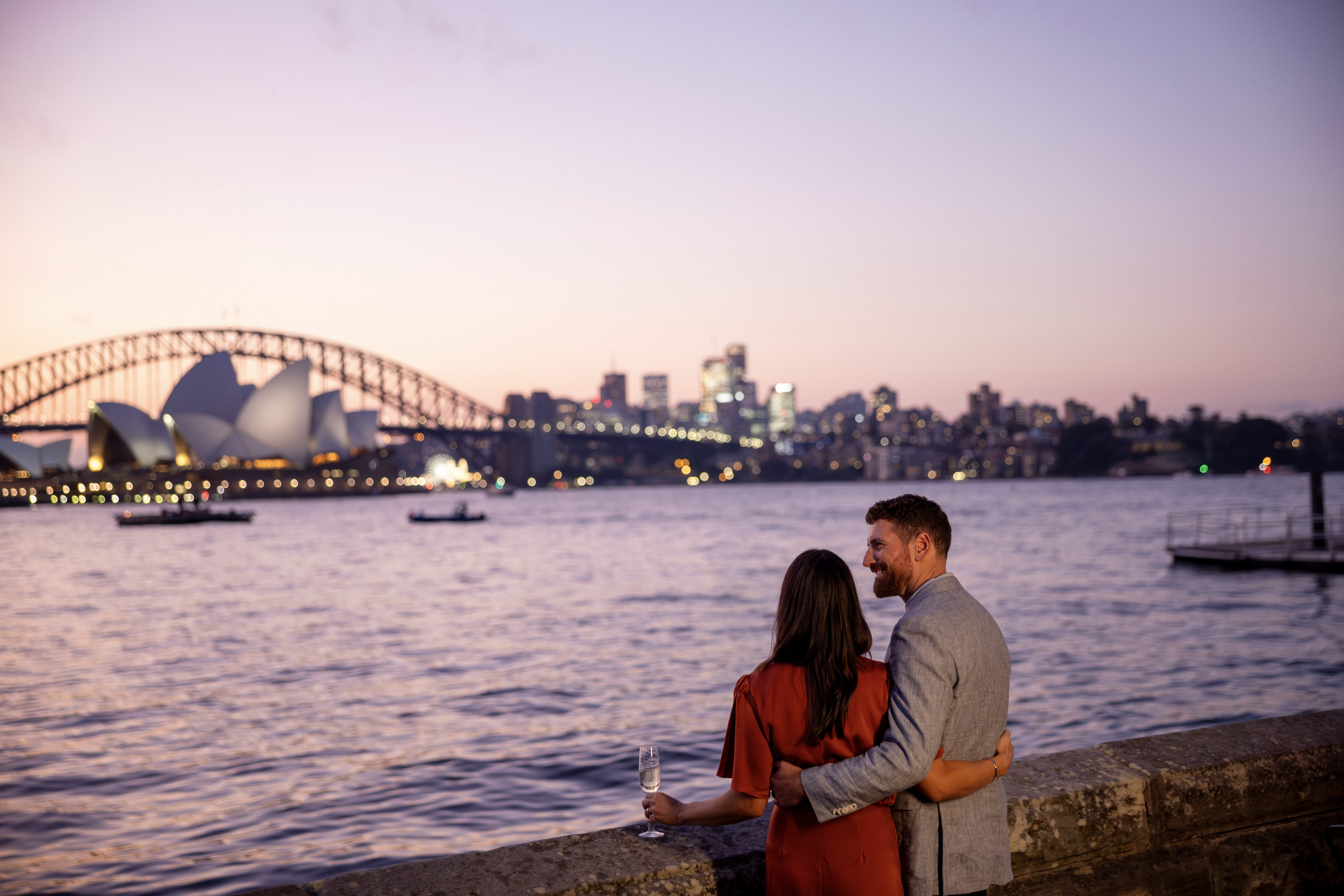 Couple arm in arm overlooking the Sydney Harbour, the Opera House and iconic bridge in the background with wine glasses in hand.