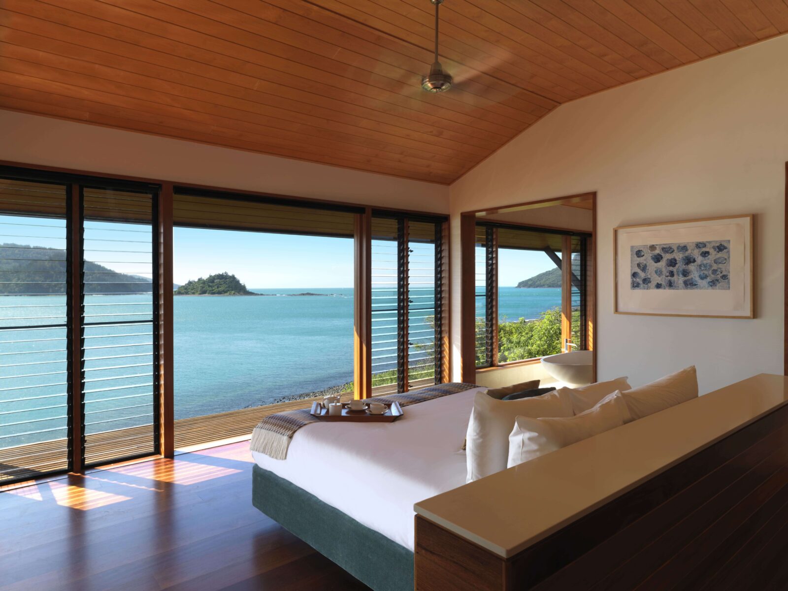 An open-plan lodge bedroom with floor-to-ceiling windows framing a turquoise bay and island views.